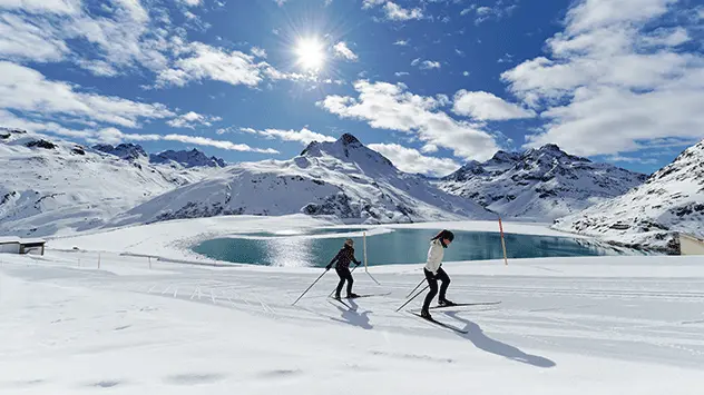 Zwei Langläufer auf einer sonnigen Piste, im Hintergrund ein See
