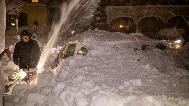 Ein Mann fährt mit einer Schneefräse zwischen parkenden Autos durch.