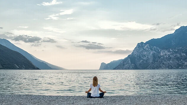 Frau sitzt im Schneidersitz am Ufer eines Sees und meditiert