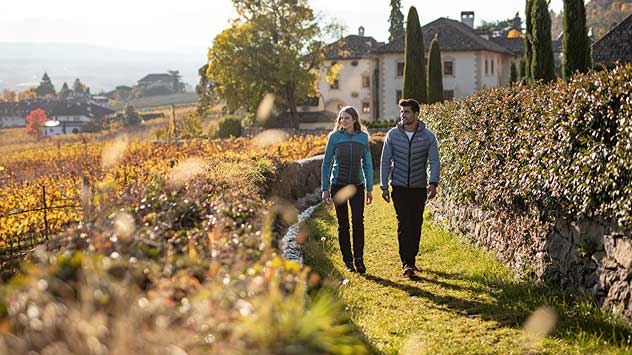 Paar spaziert auf einem Weg zwischen herbstlichen Weinbergen und Weindörfern entlang der Südtiroler Weinstraße.