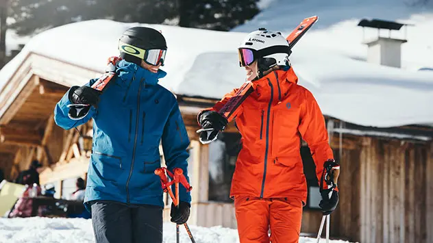 Mann und Frau tragen ihre Skier durch den Schnee zu einer Hütte.