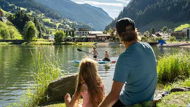 Vater und Tochter am Ufer eines Sees, mit Blick auf Kinder, die auf dem See paddeln. 