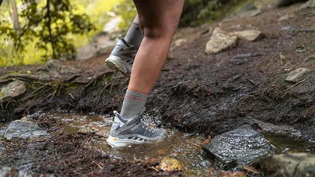 Eine Person in GORE-TEX Wanderschuhen durchquert eine Pfütze auf einem Waldweg.