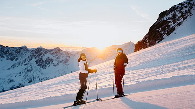 Zwei Skifahrer auf einer verschneiten Bergpiste bei Sonnenuntergang.