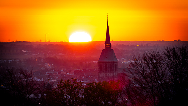 Neben dem Turm der Sankt-Andreas-Kirche in Hildesheim, dem höchsten Kirchturm Niedersachsens, verschwindet die Sonne mit gleißendem Licht am Horizont.