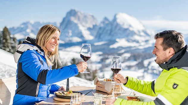 Sonnenskilauf in der Dolomitenregion Seiser Alm - Bild: IDM Südtirol / Harald Wisthaler Ein Paar stößt auf einer Skihütte mit einem Glas Wein an.