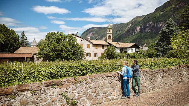 Zwei Wanderer blicken über Weinberge auf ein historisches Weindorf vor der Südtiroler Bergkulisse.