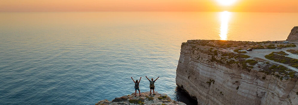 Zwei Wandernde auf einer Felsklippe an Maltas Küste bei Sonnenuntergang mit Blick auf das Meer