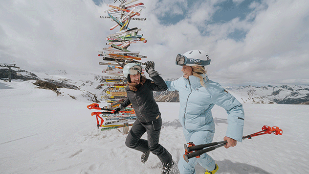 Zwei Skifahrerinnen laufen im Schnee an einem Wegweiser vorbei. 