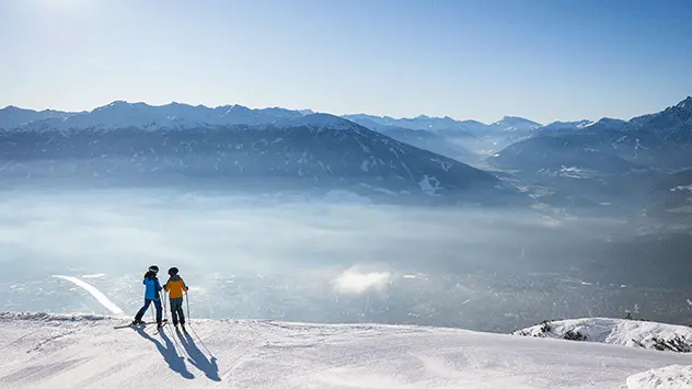 Rückansicht zweier Skifahrer, die von einem Berg aus ins Tal blicken. 