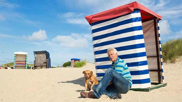 Mann sitzt mit Hund im Sand vor einem Strandkorb