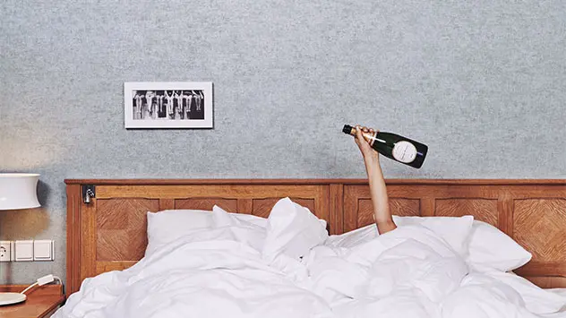 Frau liegt im Bett und hält eine Champagnerflasche in die Luft