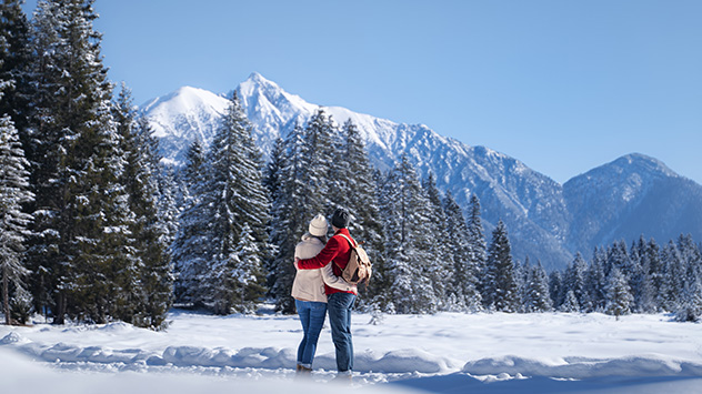 Ein Paar umarmt sich und Blickt auf die schneebedeckten Tannen sowie auf die eindrucksvolle Bergkulisse.