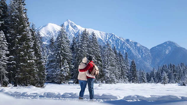 Ein Paar umarmt sich und Blickt auf die schneebedeckten Tannen sowie auf die eindrucksvolle Bergkulisse. 
