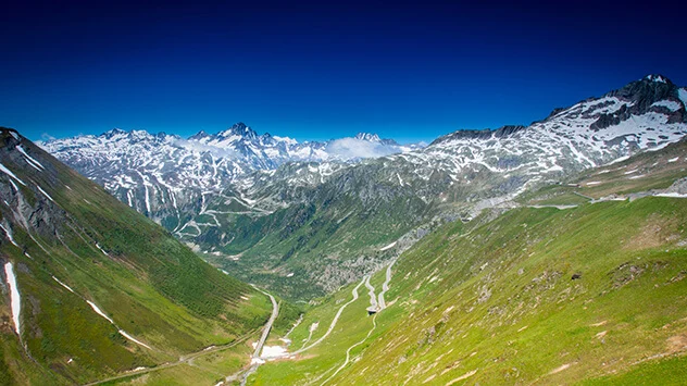 Blick über das grüne Tal und den verschneiten Furkapass am Horizont