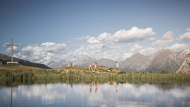 Familie auf einem Höhenweg am Bergsee, umgeben von Bergen und Wolkenhimmel.