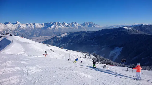 Blick über eine breite Abfahrt und darauf fahrende Skifahrer in den Bergen