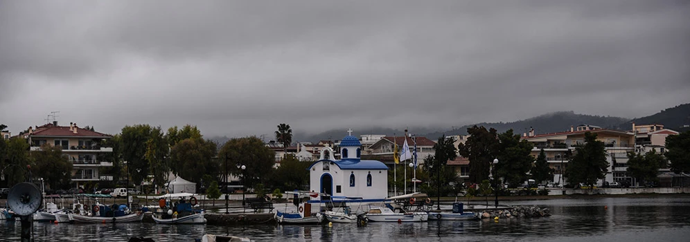 dunkle, tiefhängende Regenwolken über griechisches Dorf mit Hafen. Bewaldten Kuppen im Hintergrund