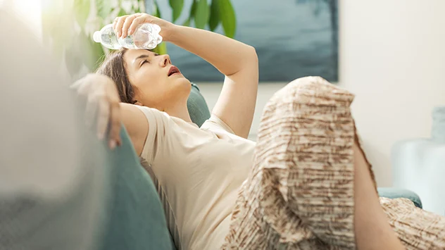 Eine Frau liegt auf dem Sofa und kühlt den Kopf mit einer Wasserflasche. 