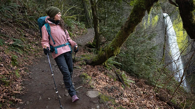 Wanderin in GORE-TEX Bekleidung auf einem Waldweg neben einem Wasserfall.