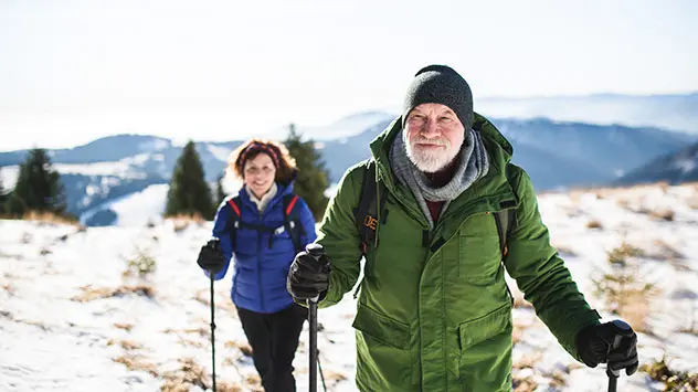 Ein Paar wandert, ausgerüstet mit Funktionskleidung und Wanderstöcken, durch die Winterlandschaft. 