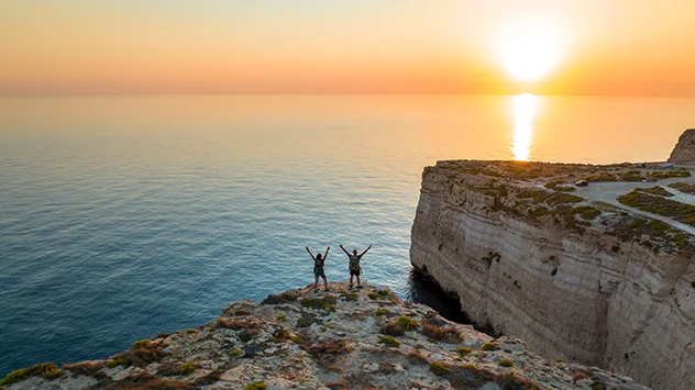 Zwei Wandernde auf einer Felsklippe an Maltas Küste bei Sonnenuntergang mit Blick auf das Meer