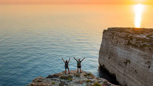 Zwei Wandernde auf einer Felsklippe an Maltas Küste bei Sonnenuntergang mit Blick auf das Meer