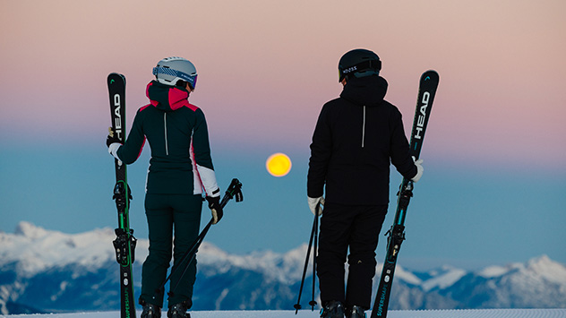 Zwei Skifahrer mit Ski in der Hand betrachten den Mond, der über den Bergen steht. 