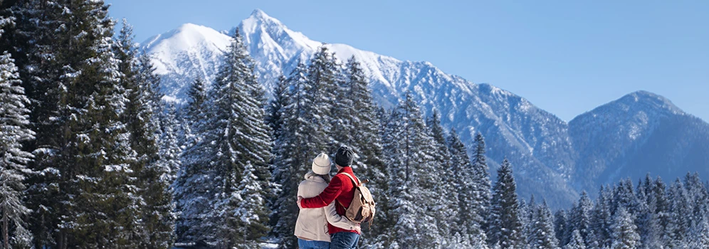 Ein Paar umarmt sich und blickt dabei auf die verschneite Winterlandschaft mit Berggipfeln. 