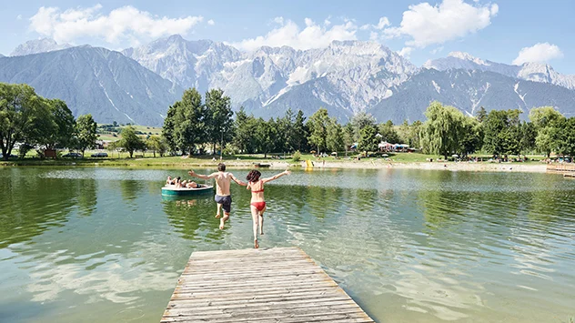 Ein Paar springt von einem Steg in einen See, im Hintergrund Bergpanorama