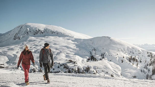 Ein Paar wandert durch die verschneite Landschaft, im Hintergrund Berge. 
