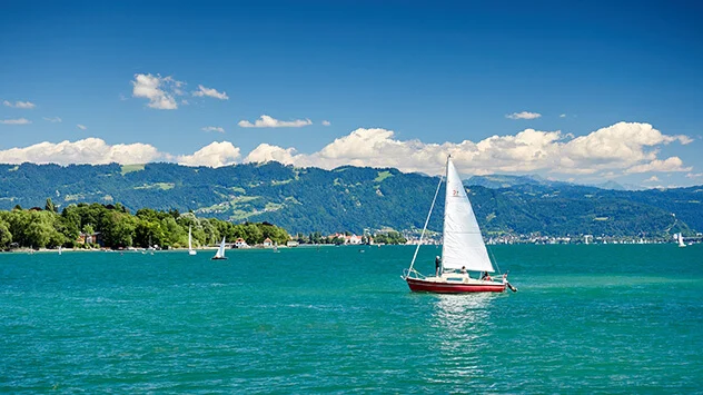 Der Boddensee mit seinem türkisen Wasser, einem Segelboot und den Alpen im Hintergrund.