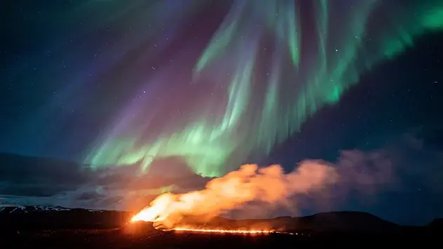 Green and purple auroras glow in the sky above the volcano, from which lava is still flowing.