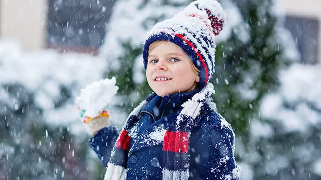 Schneefall in einem Garten - Junge mit Wollmütze und -schal hat einen Schneeball in der Hand