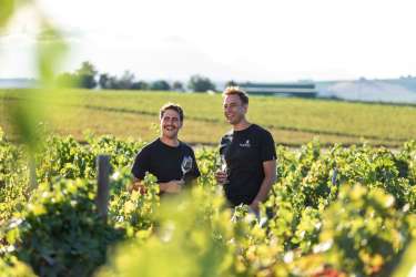 Two men standing in a vineyard holding wine glasses