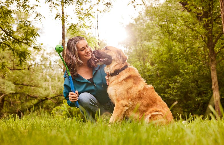 A woman in blue clothes plays with her dog using a dog toy outside on the green lawn