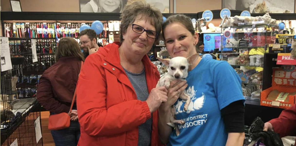 Two women holding a small white dog are standing in a store full of pet supplies