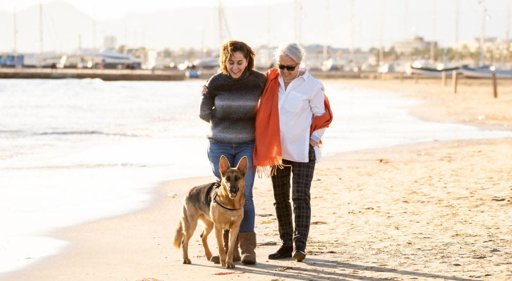 A mother and adult daughter duo laugh while walking on the beach with a large dog