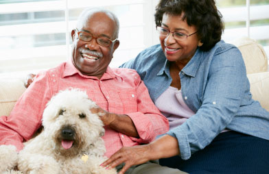 Older man and woman sitting close together and laughing while cuddling their big white dog in a home setting