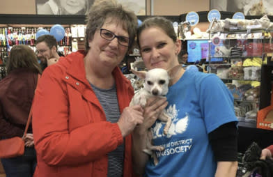 Two women holding a small white dog are standing in a store full of pet supplies