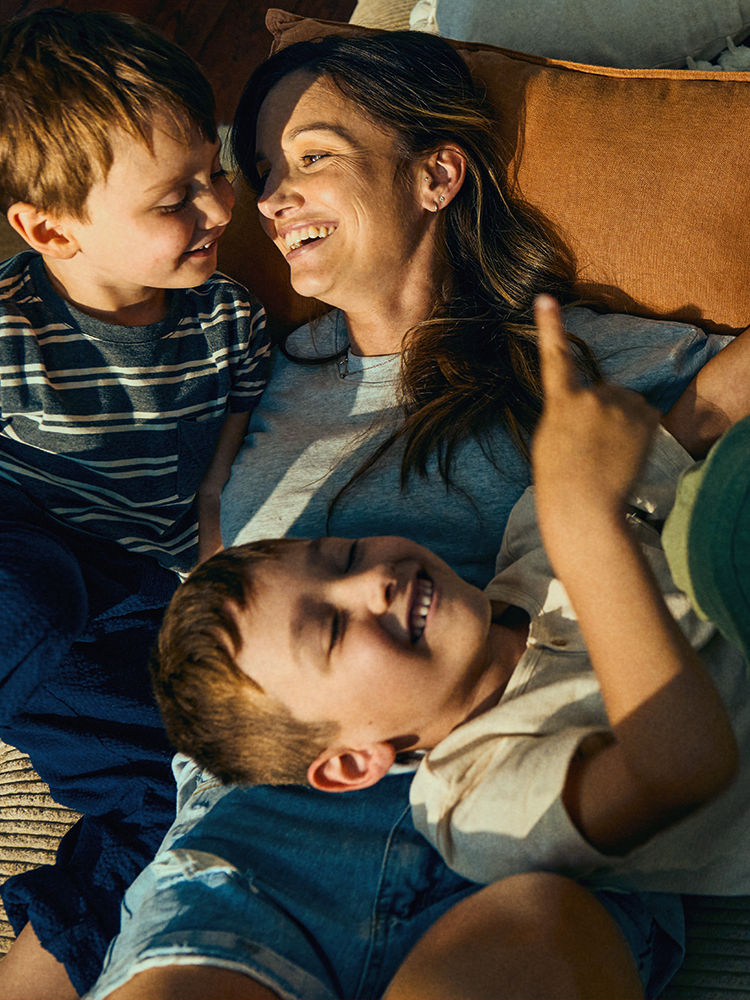 Eine Frau liegt entspannt auf einem Sofa und lacht gemeinsam mit zwei kleinen Jungen, die fröhlich neben und auf ihr liegen. Alle drei wirken ausgelassen und verbunden. 