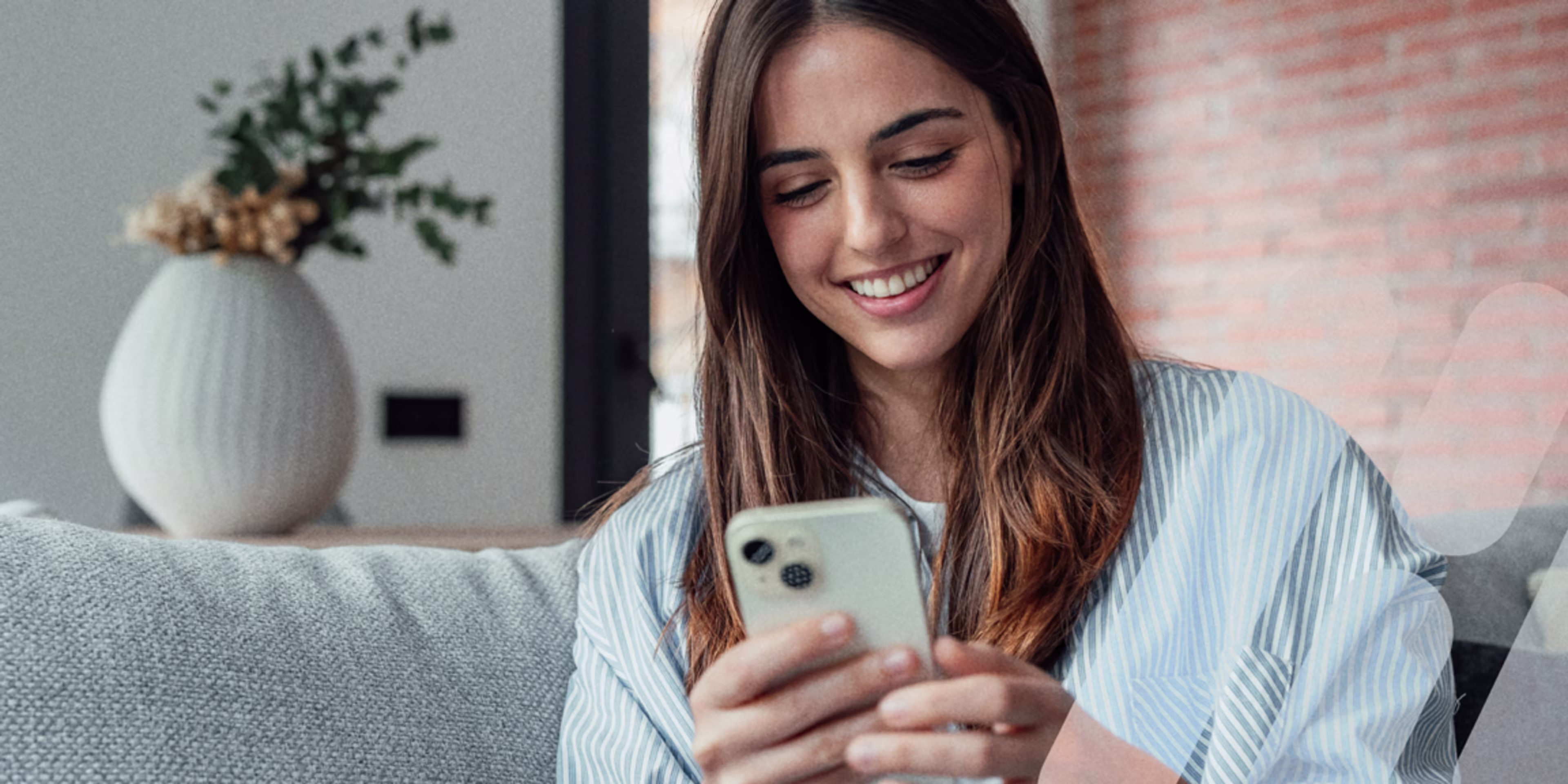 Junge Frau, sitzend, schaut lächelnd auf ein Smartphone, dass sie in ihren Händen vor sich hält.
