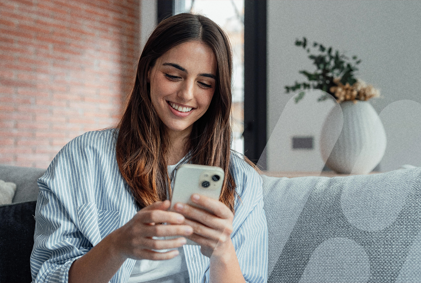 Junge Frau, sitzend, schaut lächelnd auf ein Smartphone, dass sie in ihren Händen vor sich hält.