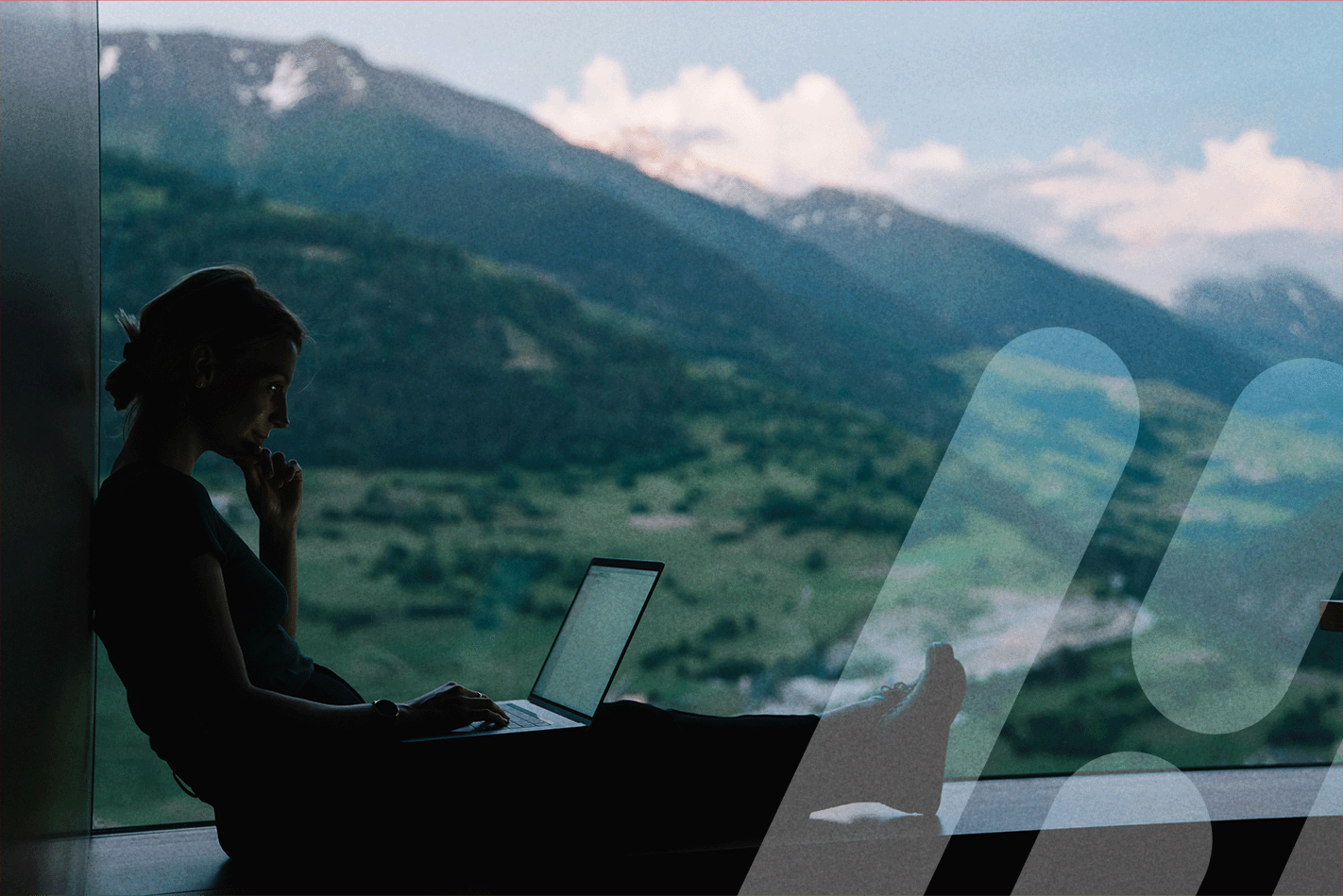 Frau sitzt am Laptop vorm Fenster mit Ausblick auf eine Berglandschaft