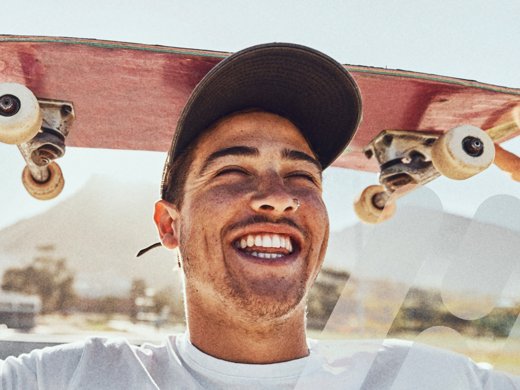 Lachender junger Mann mit Skateboard über den Schultern vor sonniger Kulisse.