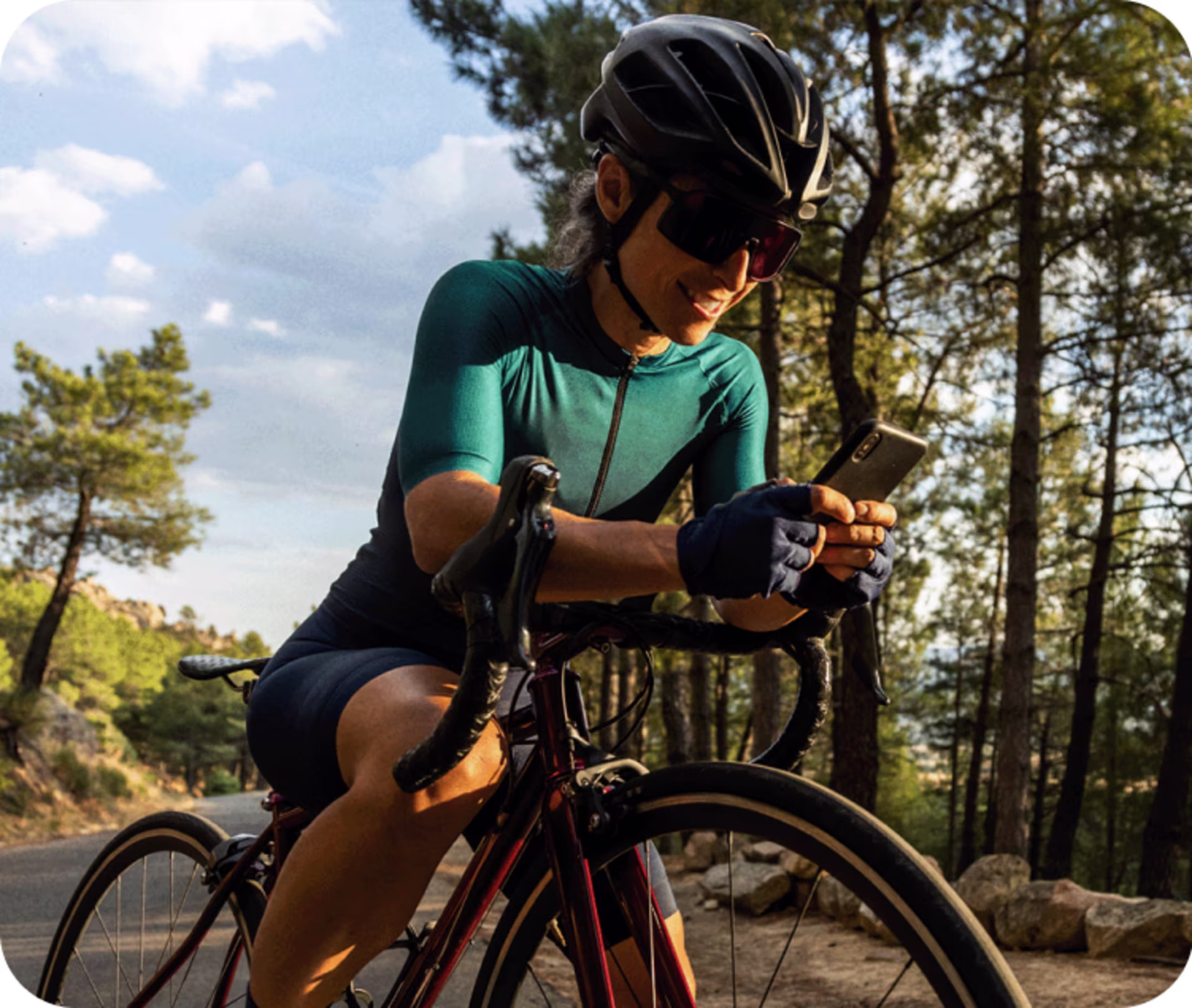 Frau in Fahrradkleidung sitzt auf Rennrad. Sie lächelt und hält auf dem Lenker gelehnt ihr Smartphone in der Hand.