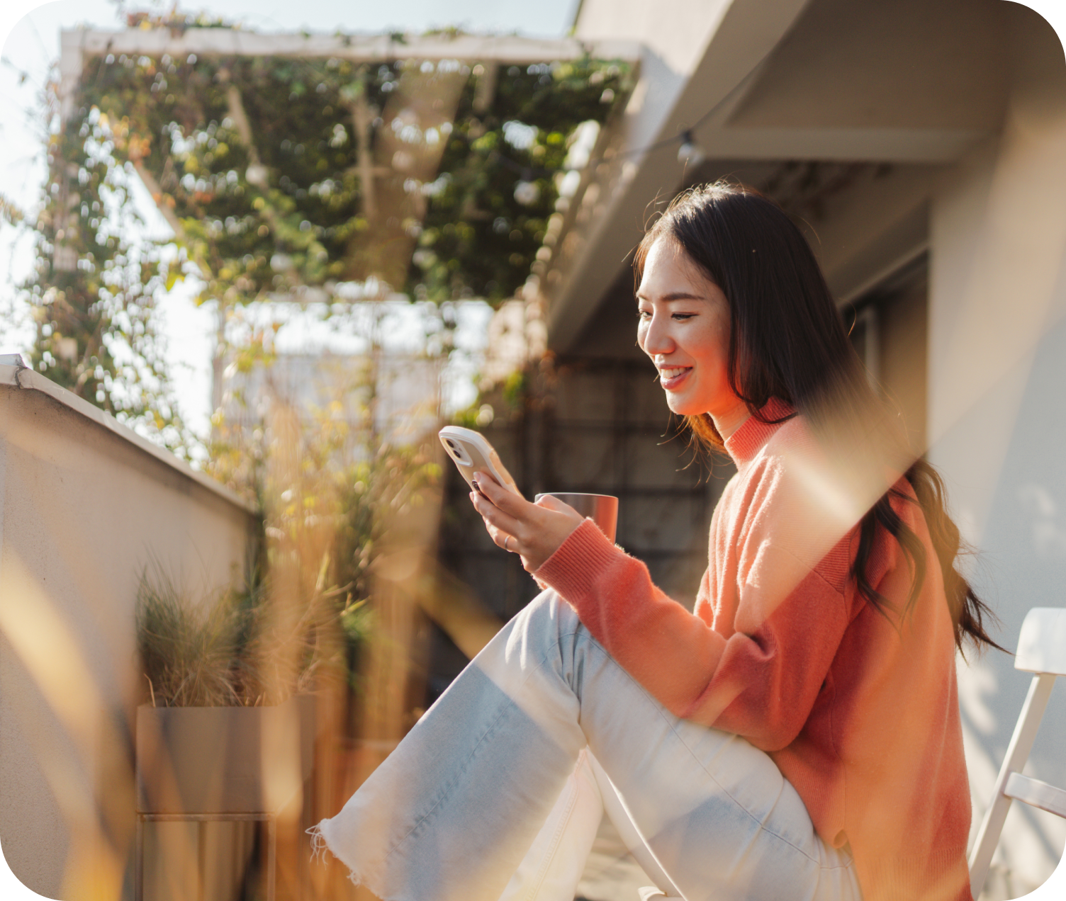 Junge Frau mit Smartphone und Kaffeetasse auf einem Balkon, lächelt beim Blick aufs Handy.