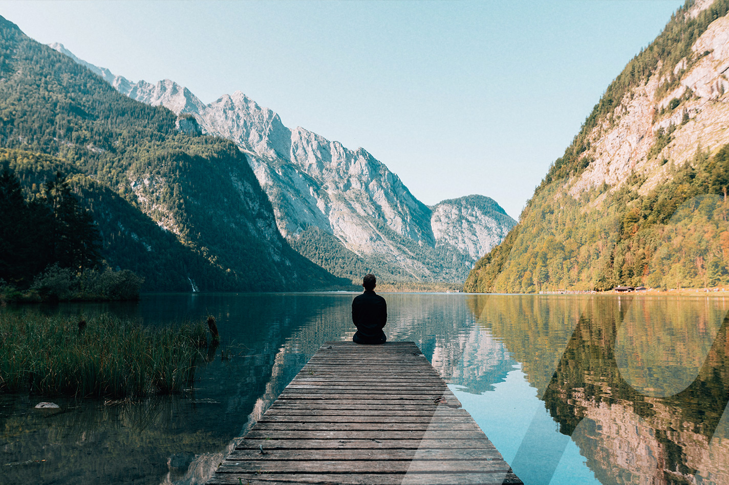Person sitzt auf einem Steg an einem Bergsee und blickt auf die umliegenden Berge.