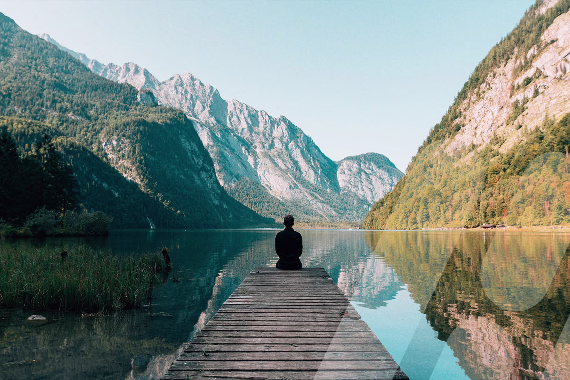 Person sitzt auf einem Steg an einem Bergsee und blickt auf die umliegenden Berge.
