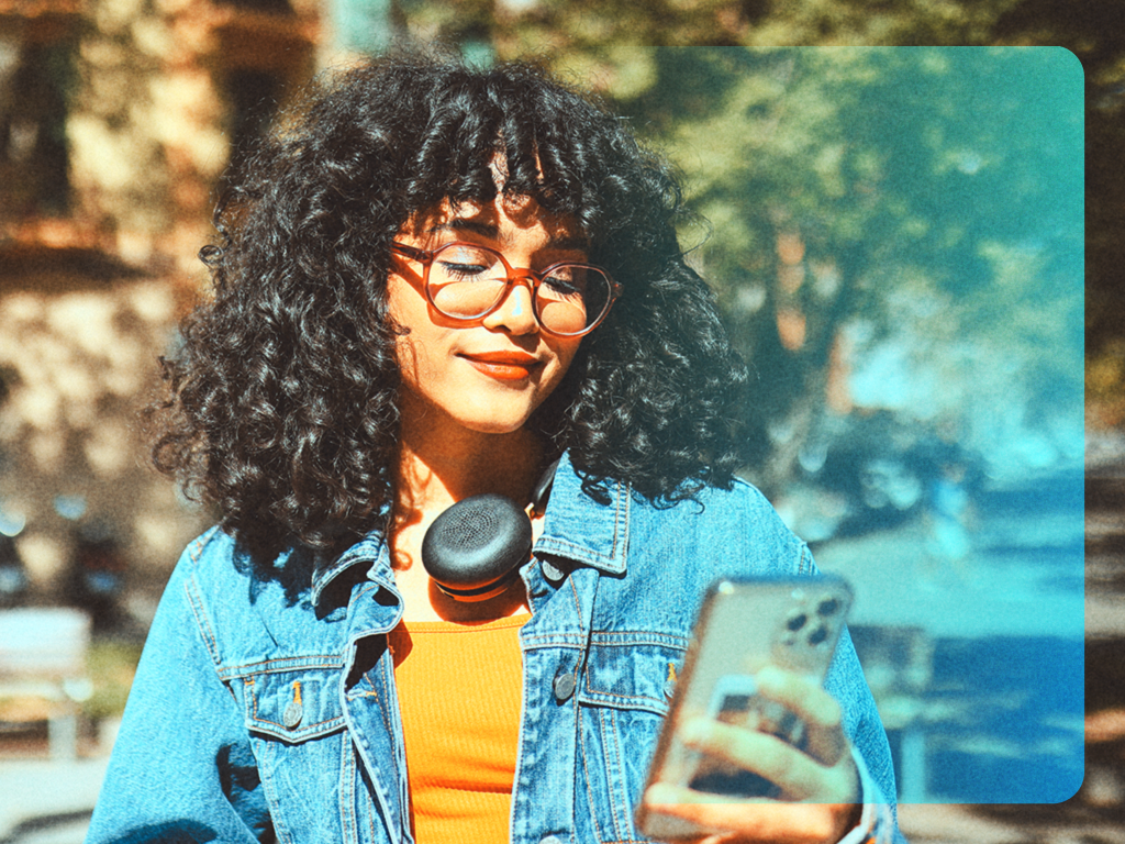 Junge Frau mit Kopfhörer um den Hals blickt auf ihr Smartphone, im Hintergrund eine sonnige Stadtstraße.